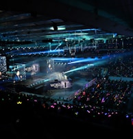 A large indoor arena filled with an audience watching a live performance. The stage features large screens displaying a performer and graphics, with bright spotlights and ambient lighting creating a dynamic atmosphere. The crowd is holding glow sticks, adding vibrant colors to the scene.