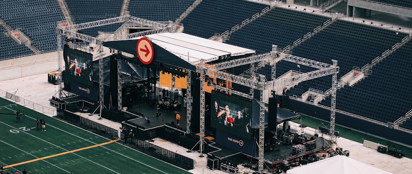 A large outdoor stage is set up inside a stadium, equipped with tall scaffolding, lights, and multiple large display screens. The seats in the stadium are mostly empty. The field is marked with numbers and lines typical of a sports venue. A few people are visible near the stage and on the field.