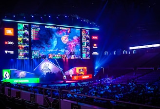 A large esports arena featuring a massive screen displaying a video game, surrounded by sponsor logos and team names. The auditorium is filled with spectators watching the gaming event. Neon blue and purple lights illuminate the stage, creating a vibrant atmosphere.
