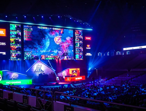 A large esports arena featuring a massive screen displaying a video game, surrounded by sponsor logos and team names. The auditorium is filled with spectators watching the gaming event. Neon blue and purple lights illuminate the stage, creating a vibrant atmosphere.