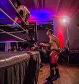 A wrestling match taking place in an indoor venue, where one participant is crouched on the top rope of the ring, while the other stands outside the ring looking at them. A crowd watches the event enthusiastically from their seats.