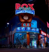 A vibrant, neon-lit facade of a gaming and sports establishment at night, featuring a large glove holding a baseball above the entrance. The building is adorned with colorful signs in both English and Japanese, with various advertisements and an illuminated sign that says 'ROX DOME'. Several people are walking towards the entrance, and bicycles are parked along the side.