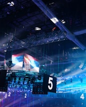 A large indoor arena is equipped with advanced lighting and audio systems. Suspended above the space are numerous digital displays showcasing numbers. The environment is filled with blue illumination and spotlights creating a vibrant atmosphere.