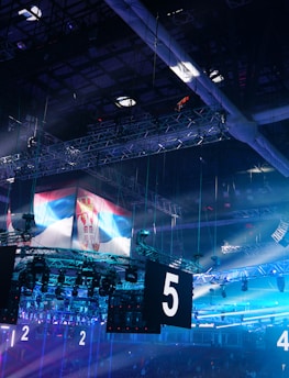 A large indoor arena is equipped with advanced lighting and audio systems. Suspended above the space are numerous digital displays showcasing numbers. The environment is filled with blue illumination and spotlights creating a vibrant atmosphere.