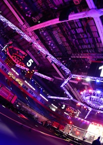 An indoor arena with vibrant purple and blue lighting, featuring large numbered screens suspended from the ceiling. The venue has a high ceiling with intricate metal truss structures and hanging lights, creating a futuristic and energetic atmosphere.