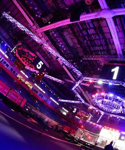 An indoor arena with vibrant purple and blue lighting, featuring large numbered screens suspended from the ceiling. The venue has a high ceiling with intricate metal truss structures and hanging lights, creating a futuristic and energetic atmosphere.