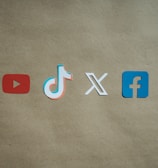 Four social media platform logos are arranged on a brown background. From left to right, the logos are for YouTube in red, TikTok in a combination of red, blue, and white, X in white, and Facebook in blue.