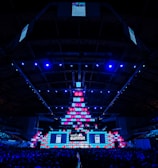 An expansive venue filled with a large audience, featuring a grand stage lit with vibrant blue and pink lights. The ceiling is decorated with a large display, and numerous screens showcase the event's branding. The atmosphere is dynamic and futuristic, reflecting a technologically advanced conference.