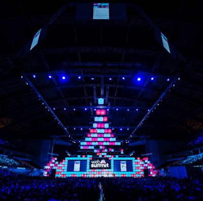 An expansive venue filled with a large audience, featuring a grand stage lit with vibrant blue and pink lights. The ceiling is decorated with a large display, and numerous screens showcase the event's branding. The atmosphere is dynamic and futuristic, reflecting a technologically advanced conference.